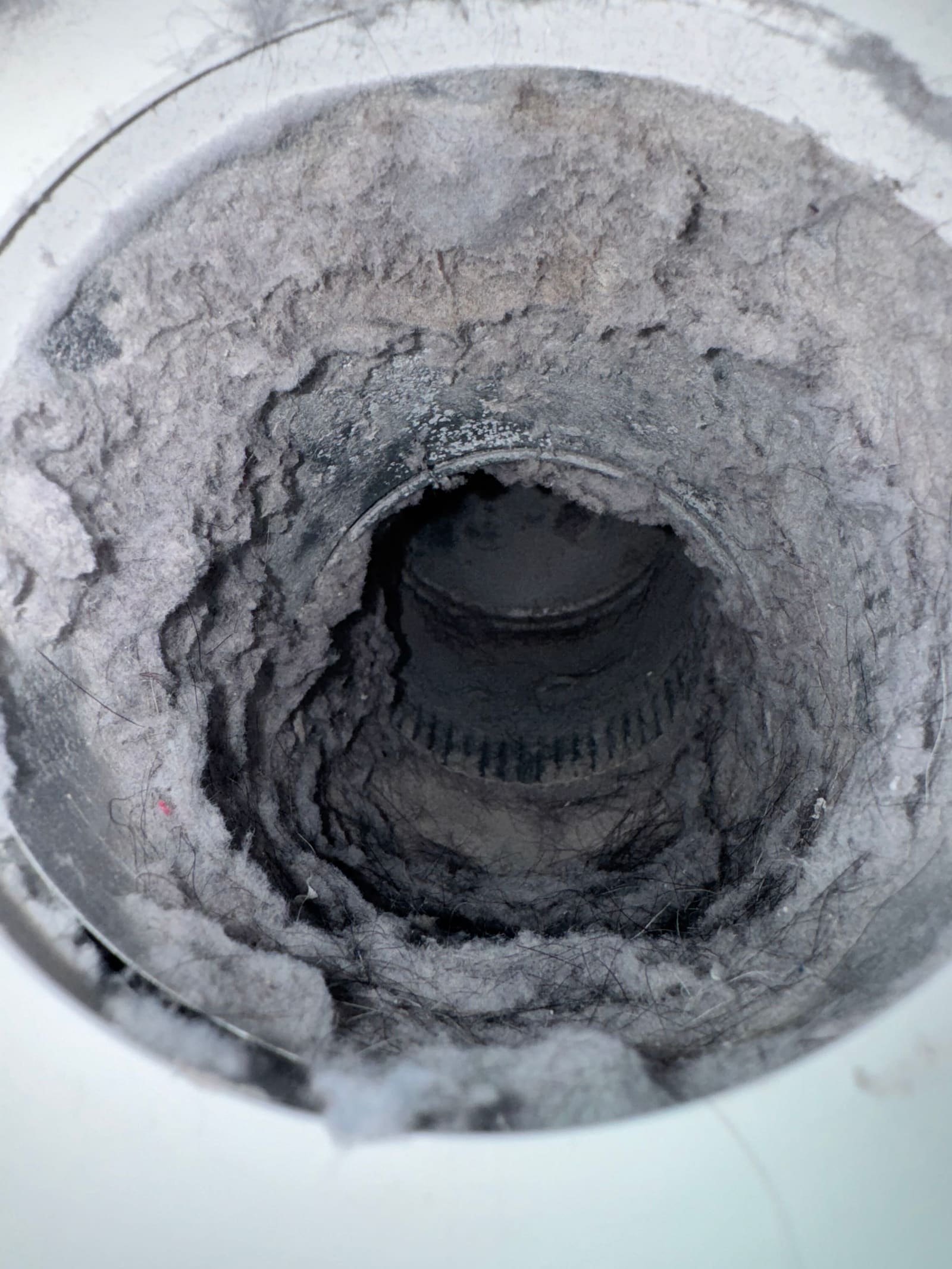 View down a dryer vent duct where compacted grey lint has narrowed the passage to a small dark opening.